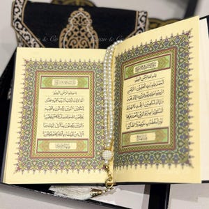 May include: An open Quran with a white prayer bead necklace resting on top of the pages. The pages are decorated with intricate gold and green designs. The book is resting on a black surface.
