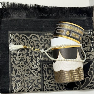 May include: A black prayer rug with a gold and silver patterned design, a white prayer bead necklace, and a black and gold cylindrical container with the text "Azan & Co" on the side.