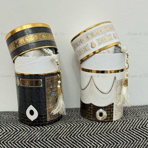 以下が含まれることがあります： 金色の縁取りとイスラムのデザインが施された装飾的な容器が2つ。容器は黒と白で、金色の房飾り付き。容器には「Azaan & Co.」のラベルが貼られています。