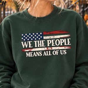 May include: A forest green crewneck sweatshirt featuring a distressed American flag graphic. The text reads WE THE PEOPLE in large letters, with MEANS ALL OF US printed below in a smaller font. The design has a weathered, vintage appearance.