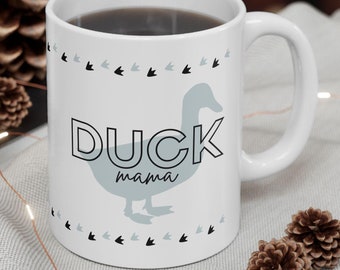 May include: White ceramic mug with the words "DUCK mama" and a light blue duck illustration. The mug has a black and gray border of duck footprints. The mug is filled with a dark liquid.