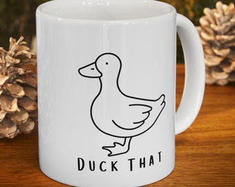 May include: White ceramic mug with a black outline drawing of a duck and the words "DUCK THAT" in black. The mug has a curved handle and is set on a wooden surface with pine cones in the background.