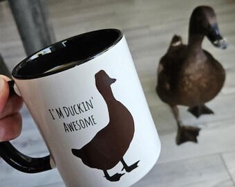 May include: White ceramic mug with a black handle and rim, featuring a brown duck silhouette and the text "I'M DUCKIN' AWESOME." A real duck is in the background. The mug is held by a person.