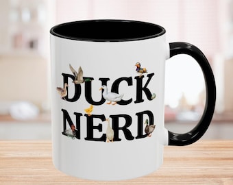 May include: White ceramic coffee mug with a black rim and handle. The mug has a black and white graphic of ducks and the text "Duck Nerd".