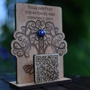 Könnte beinhalten: Ein dekoratives Holzstück mit einem Baumdesign und dem deutschen Text "FOLGE DEM PFAD UND ENTDECKE, WAS VERBORGEN LIEGT." Eine blau-weiße Glasperle ist befestigt. Ein kleiner QR-Code aus Holz ist ebenfalls vorhanden.