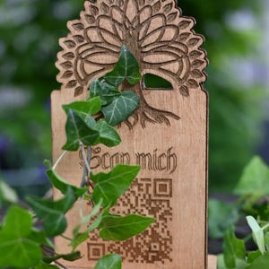 Könnte beinhalten: Ein Holzschild mit einem Baumdesign und dem Schriftzug "Scan mich" sowie einem QR-Code. Grüne Efeublätter sind über das Schild drapiert. Das Schild ist aus hellem Holz gefertigt. Ideal für ein Geschäft.