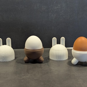 May include: Four white ceramic egg holders shaped like bunnies, with two holding white eggs and one holding a brown egg.