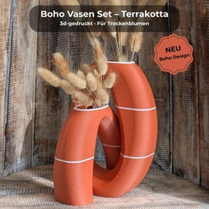 Pode incluir: Um conjunto de vasos cor de terracota com um design único e entrelaçado. Os vasos têm listras brancas e contêm flores secas. A imagem inclui o texto "Boho Vasen Set - Terrakotta" e "NEU Boho Design."
