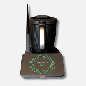 May include: A black kitchen appliance with a vertical handle and a tall, thin accessory. The appliance sits on a brown base with the text "Sabrina's TM7" in a green circular design. The appliance has a ribbed texture.
