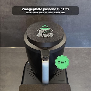 May include: A black Thermomix TM7 scale cover plate with a green and white graphic of a cauldron. The cover plate is on top of a black Thermomix appliance with a silver handle. The text "2 in 1" is on a green sticker.