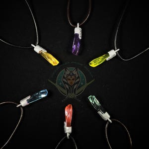 Peut inclure: Collection de sept colliers pendentifs en cristal de différentes couleurs, dont le jaune, le violet, le bleu, le rouge et le vert. Chaque pendentif est attaché à un fermoir en métal argenté et à un cordon fin. Les colliers sont disposés sur une surface noire.