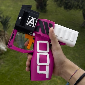 May include: A magenta and black toy gun with the word "DOT" in white. The gun has an orange trigger and a white barrel. The letter "A" is visible on the black top of the gun.