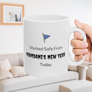 May include: White ceramic mug with a handle, featuring the text "Marked Safe From Mamdani's New York Today" in black font. A blue flag graphic is above the text. The mug is held by a person.