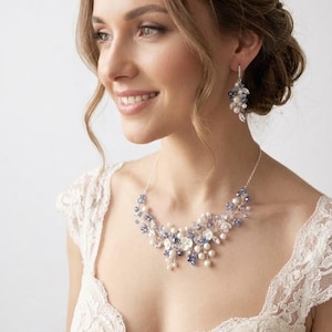 May include: A silver-toned necklace and matching drop earring set featuring clusters of white pearls, clear crystals, and small blue beads arranged in a floral-inspired pattern on a delicate metal chain with a secure clasp closure.