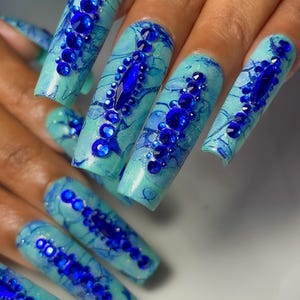 May include: Long, light blue, almond-shaped acrylic nails with a marbled design and blue rhinestones. The nails are decorated with a variety of blue rhinestones, including small round stones and larger, rectangular stones. The text "Nails By Aquarian Legs" is visible in the bottom right corner of the image.