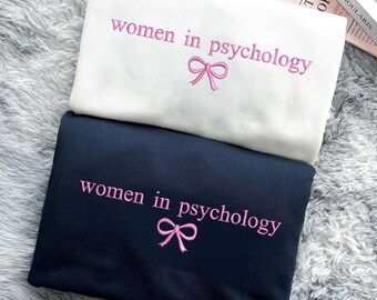 May include: Two sweatshirts with the text "women in psychology" and a pink bow embroidered on each. The top sweatshirt is white and the bottom sweatshirt is black.