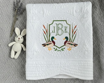May include: White quilted baby blanket with an embroidered duck design. The blanket features a central emblem with the initials "JBE" and two ducks. A small crocheted bunny and lavender sprig are next to the blanket.