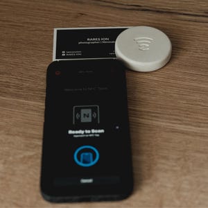 Peut inclure: Un smartphone avec un écran noir affichant "Ready to Scan" et une icône circulaire. Une étiquette NFC blanche et ronde avec un symbole Wi-Fi est à côté d'une carte de visite qui dit "RARES ION photographe | cinéaste".