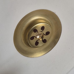 Peut inclure: Gros plan d'un évier en porcelaine blanche avec un bouchon de vidange en laiton. Le bouchon de vidange a un motif décoratif de six petits cercles brun foncé.