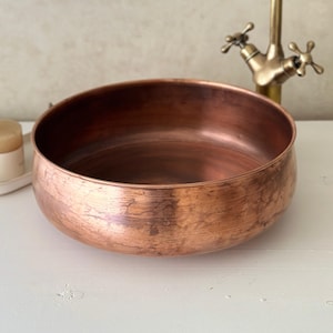 May include: A copper-colored, round basin with a brushed finish. A vintage-style faucet is in the background. A small candle in a white holder sits to the left. The basin is likely for a bathroom or kitchen.