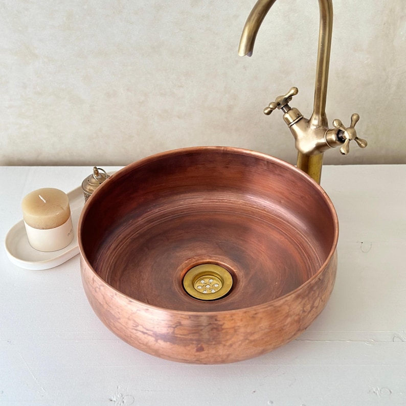May include: A round copper vessel sink with a brass drain and a brass faucet with two handles. The sink is on a white countertop.