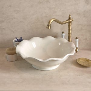 May include: A white ceramic sink with a scalloped edge, paired with a brass tap and white-handled fixtures. A small gold soap dish and a candle are on the counter. The sink has a decorative, vintage style.