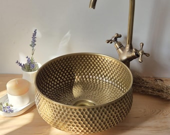 May include: A hammered brass vessel sink with a matching faucet. The sink has a textured, dimpled surface and a round shape. The faucet has a curved spout and cross handles. A small white vase with lavender and a candle are on the wooden surface.