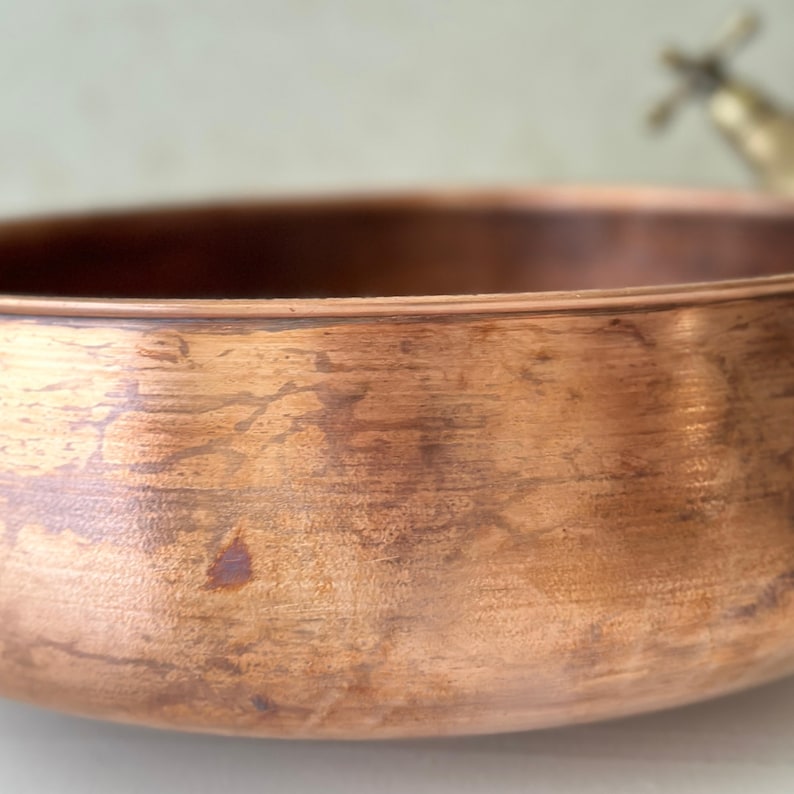 May include: Close-up of a copper bowl with a textured surface and a warm, reddish-brown hue. The bowl has a rounded shape and a slightly flared rim. The metal has a natural, aged appearance, suggesting it is handmade.