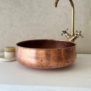 May include: A copper-colored, round basin with a textured surface. A vintage-style faucet is visible in the background. A small candle in a white holder sits to the left. The basin is a decorative element for a bathroom.