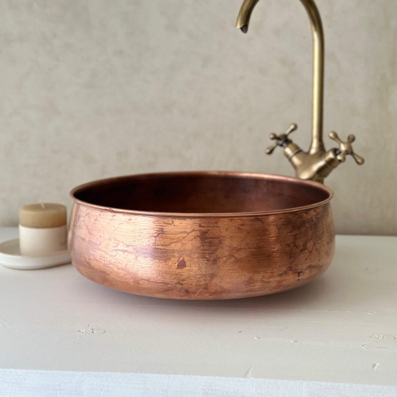 May include: A copper-colored, round basin with a textured surface. A vintage-style faucet is visible in the background. A small candle in a white holder sits to the left. The basin is a decorative element for a bathroom.