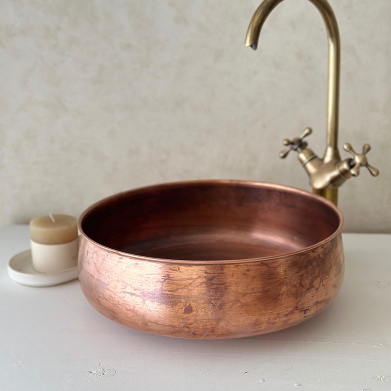 May include: A copper-colored, round basin with a textured surface. A vintage-style brass faucet is positioned behind the basin. A small candle in a white holder sits to the left. The items are on a white surface.