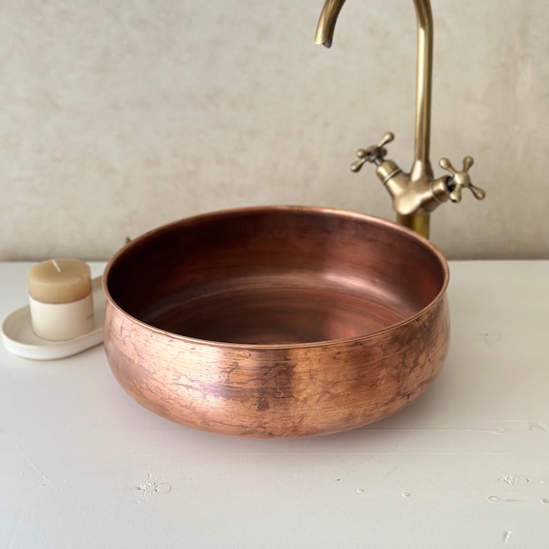 May include: A copper-colored, round basin with a brushed finish. A vintage-style faucet is in the background. A small candle in a white holder sits to the left. The basin is likely for a bathroom or kitchen.