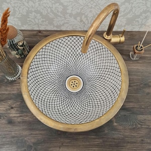 Peut inclure: Un lavabo en céramique blanche avec un motif géométrique noir et une bordure dorée. Le lavabo est rond et possède un drain au centre. Un robinet doré est visible en arrière-plan.