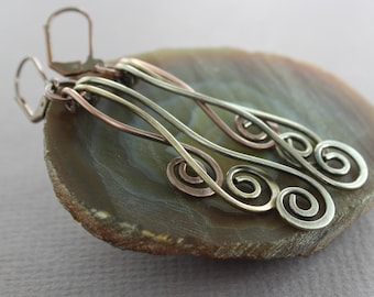 May include: A pair of long, silver and copper wire earrings with multiple spiral designs. The earrings are displayed on a brown agate slice.