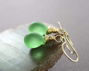 May include: A pair of green glass teardrop earrings with gold wire wrapped around the top of each teardrop. The earrings are hanging from gold hooks.