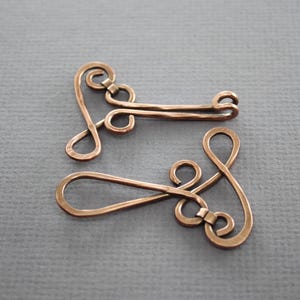 May include: Two copper wire clasps with a decorative swirl design. The clasps are made of a single piece of wire that is bent and twisted to create the design.