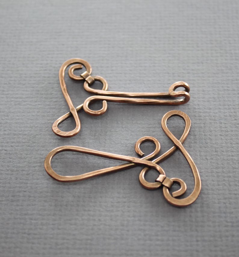 May include: Two copper wire clasps with a decorative swirl design. The clasps are made of a single piece of wire that is bent and twisted to create the design.