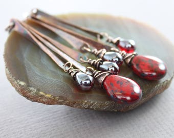 May include: A set of four long, copper earrings with red and silver teardrop-shaped beads. The earrings are displayed on a brown agate slice.