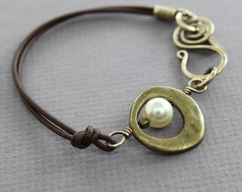 May include: A brown leather bracelet with a brass charm featuring a circle with a white pearl in the centre. The charm is attached to a twisted wire design.