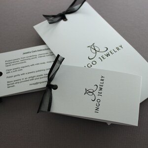 May include: Two white cards with black text and a black ribbon. The top card has the text "Jewelry Care Instructions" and instructions on how to care for jewelry. The bottom card has the text "Ingo Jewelry" in a stylized font.