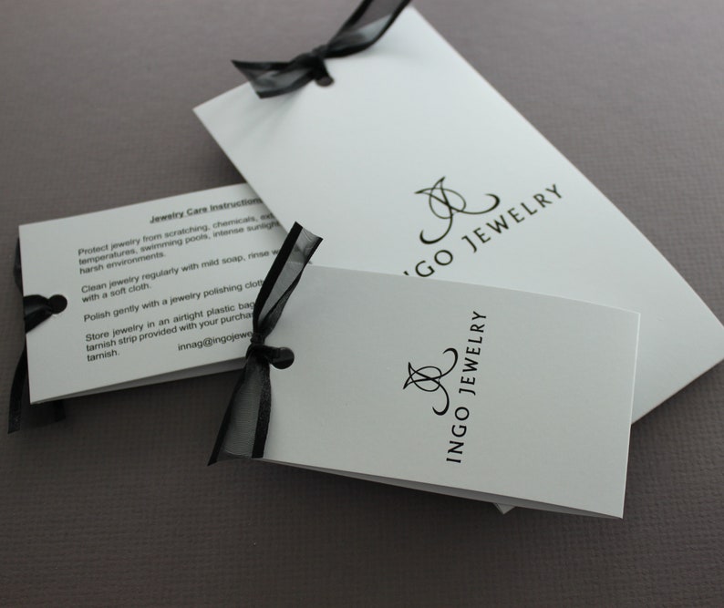 May include: Two white cards with black text and a black ribbon. The top card has the text "Jewelry Care Instructions" and instructions on how to care for jewelry. The bottom card has the text "Ingo Jewelry" in a stylized font.