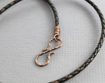 May include: A braided leather cord necklace with a copper clasp. The cord is black and brown and has a woven pattern. The clasp is a simple hook and eye design.