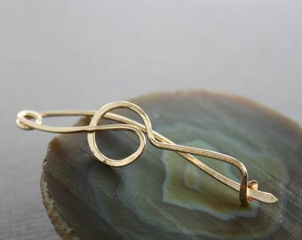 May include: A gold-toned metal hair clip with a circular design. The clip is on a brown and grey background.