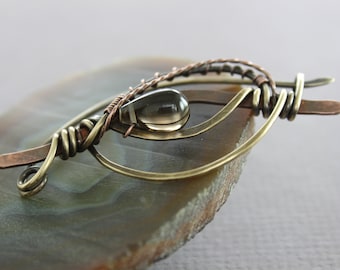 May include: A smoky quartz gemstone is set in a wire wrapped pendant with copper and brass wire. The pendant is displayed on a slice of agate.