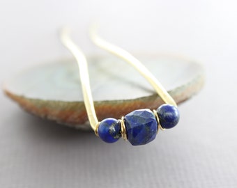 May include: A gold-coloured hair pin featuring three dark blue lapis lazuli beads. The central bead is a faceted, geometric shape, with two round beads either side. The pin has a simple, elegant, curved design.