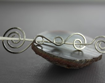 May include: Silver hair stick with a swirling design. The stick is resting on a gray and brown agate stone.