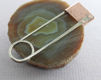 May include: A silver safety pin with a hammered finish and a copper accent. The pin is resting on a slice of agate with brown and white banding.