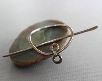 May include: A copper hair stick with a circular design and a knot at the end. The stick is resting on a piece of agate.