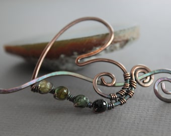 May include: A handcrafted copper-toned metal hair accessory in a heart shape, adorned with green and black beads. The design features a swirling, decorative element. The background is blurred.
