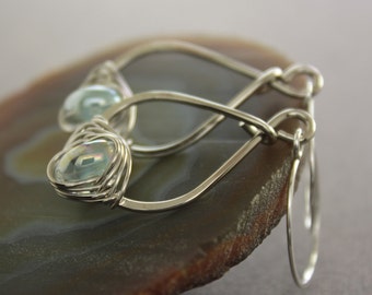 May include: Silver wire wrapped earrings with iridescent blue gemstone accents. The earrings are teardrop shaped and dangle from silver hooks.